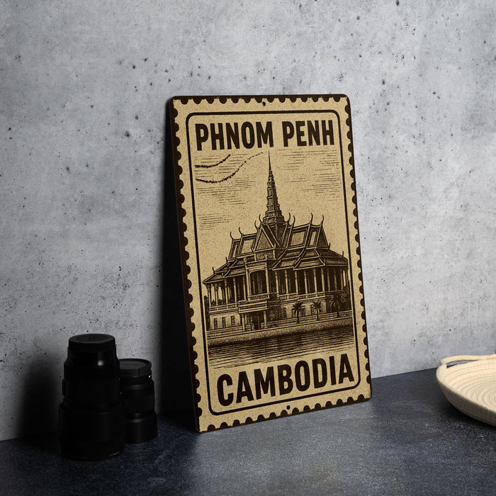 A metal sign with the text "PHNOM PENH" and "CAMBODIA" printed on it, along with an illustration of a temple or building. The sign is placed on a table next to some black camera lenses.