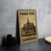 A metal sign with the text "PHNOM PENH" and "CAMBODIA" printed on it, along with an illustration of a temple or building. The sign is placed on a table next to some black camera lenses.