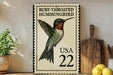 A framed postage stamp featuring a hummingbird, with the text "Ruby-throated Hummingbird" and "USA 22" printed on it. The stamp is placed on a wooden surface, with a potted plant, a cutting board, and some lemons nearby.