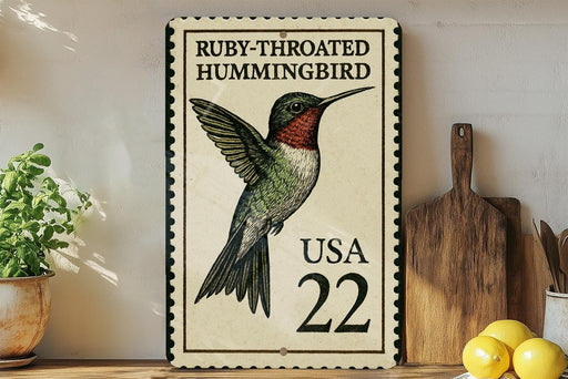 A framed postage stamp featuring a hummingbird, with the text "Ruby-throated Hummingbird" and "USA 22" printed on it. The stamp is placed on a wooden surface, with a potted plant, a cutting board, and some lemons nearby.