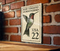 A framed image of a hummingbird, which appears to be a postage stamp, is mounted on a brick wall.