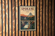 A vintage-style postage stamp depicting a scenic view of Spokane, Washington, USA, with a bridge and waterfall in the background.