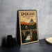 A framed poster of the Spokane, Washington, USA, featuring a scenic illustration of a bridge, waterfall, and mountains.