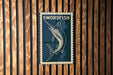 A metal sign with the word "SWORDFISH" printed on it, mounted on a wooden wall.