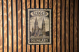 A vintage-style postage stamp from Hungary, featuring a church with two towers, is displayed on a wooden wall.