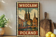 A vintage-style metal sign with a painting of a cityscape and the text "Wrocław Poland" displayed on it, placed on a shelf next to a wooden cutting board and some potted plants.