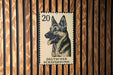 A German Shepherd dog is depicted on a German postage stamp, which is affixed to a wooden wall.