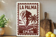 a framed poster of La Palma, Spain, with a palm tree and a mountain in the background. The poster is displayed on a shelf next to a wooden cutting board and a potted plant.