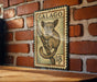 A vintage postage stamp featuring a cartoon illustration of a small animal, likely a koala, is displayed on a brick wall.
