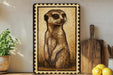 a framed illustration of a meerkat, a small burrowing mammal, on a shelf next to a wooden cutting board and a potted plant.