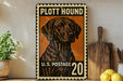 A vintage-style stamp featuring a hound dog, with the text "PLOTT HOUND" and "U.S. POSTAGE 20" printed on it. The stamp is displayed on a shelf next to a wooden cutting board and a potted plant.