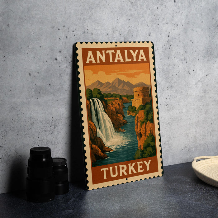 A vintage-style metal sign with the text "ANTALYA TURKEY" and an illustration of a waterfall and mountains, placed on a gray countertop next to some black camera lenses.