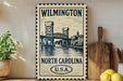 A vintage-style metal sign with a drawing of a bridge and the text "Wilmington North Carolina U.S.A." displayed on a wooden shelf.