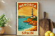 A vintage-style metal stamp depicting a lighthouse and a coastal landscape, with the text "MENORCA SPAIN" on it, is displayed on a shelf next to a potted plant and a wooden cutting board.