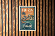 A vintage-style travel poster for Ibiza, Spain, featuring a tropical beach scene with palm trees and buildings.