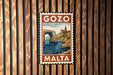 A vintage-style Malta postage stamp is displayed on a wooden wall.