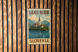 A vintage-style postage stamp depicting Lake Bled, a famous landmark in Slovenia, is displayed on a wooden wall.