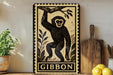 a framed illustration of a monkey, with the word "GIBBON" written below it. The monkey is depicted in a playful pose, with its arms raised above its head. The frame is placed on a wooden surface, with a potted plant and a cutting board nearby.