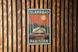 A vintage-style metal sign with the text "ISLAMABAD PAKISTAN" displayed on a wooden wall.