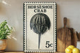 A metal sign with a black and white illustration of a horseshoe crab, mounted on a wall next to a wooden cutting board and a potted plant.
