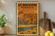 a vintage-style poster of Ait Benhaddou, Morocco, displayed on a wall next to a wooden cutting board and some potted plants.