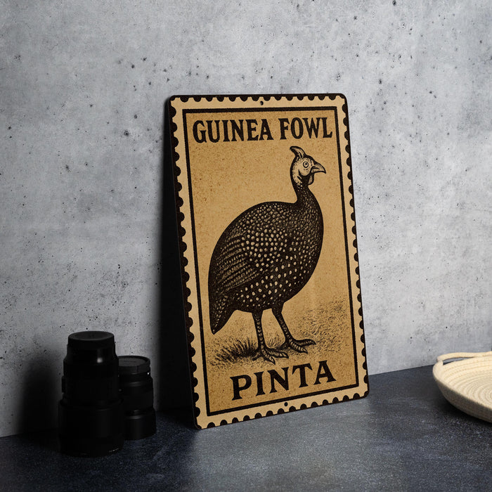 A metal sign with a printed image of a guinea fowl and the text "GUINEA FOWL PINTA" is displayed on a table.