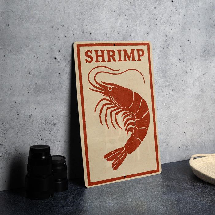 A sign with the word "SHRIMP" on it, featuring a red shrimp illustration, is placed on a countertop next to some black containers.