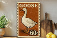 A vintage-style metal sign featuring a cartoon goose, with the text "GOOSE 60c" printed on it. The sign is displayed on a shelf next to a wooden cutting board and a potted plant.