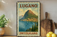 A vintage-style poster of Lugano, Switzerland, featuring a mountain landscape with a town nestled at the base. The poster is framed and displayed on a shelf, with a potted plant, a wooden cutting board, and some lemons nearby.