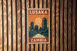 A vintage-style postage stamp from Zambia, featuring a cityscape with tall buildings and a sunset in the background.