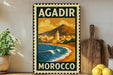 A vintage-style metal stamp with the text "AGADIR MOROCCO" and an illustration of a coastal city with mountains in the background.