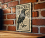 a wooden table with a framed illustration of a black bird, which appears to be a stamp or a sign with the word "GRACKLE" written on it.
