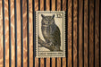 A postage stamp featuring a great horned owl is displayed against a wooden background.