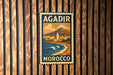 A vintage-style metal stamp with the text "AGADIR MOROCCO" is displayed on a wooden wall.