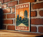 A vintage-style metal sign with the text "LUSAKA ZAMBIA" displayed on a brick wall.