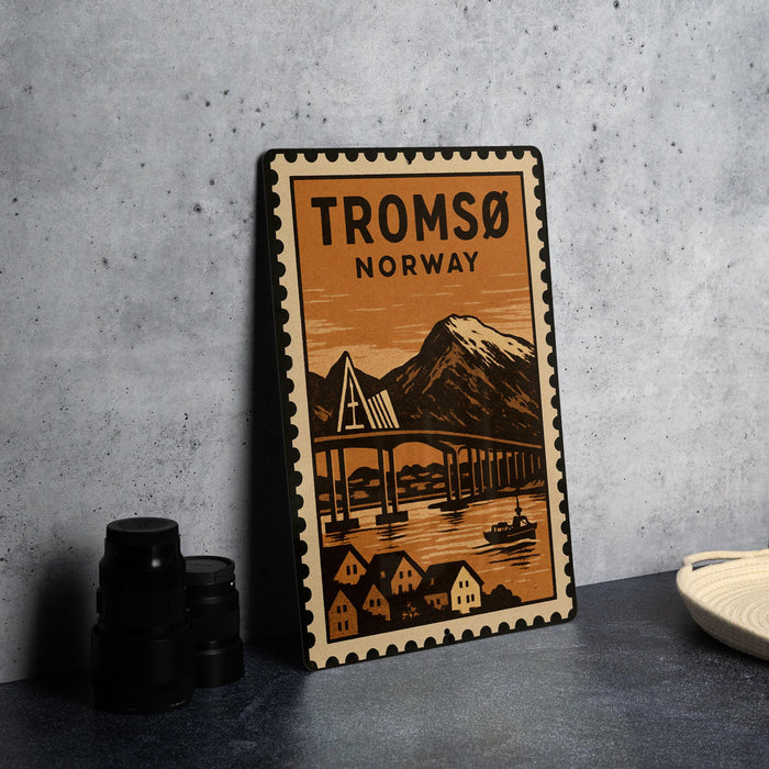 A metal poster with a vintage-style illustration of a bridge and mountains, along with the text "TROMSO NORWAY" printed on it, is displayed on a table.