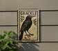 a black bird with a stamp-like border, mounted on a wall next to a plant.