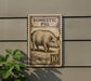 a sign that reads "Domestic Pig" and has a stamp-like design, mounted on a wall next to a plant.
