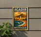 A vintage-style metal sign with the text "AGADIR MOROCCO" and an illustration of a coastal city with a mountain in the background.