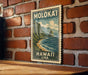 A vintage-style postage stamp depicting a mountainous landscape with a beach and ocean, hanging on a brick wall.