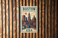 A vintage-style poster of Boston, Massachusetts, featuring a cityscape with buildings, a clock tower, and a bridge, is displayed on a wooden wall.