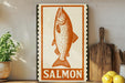 a framed illustration of a salmon fish, with the word "SALMON" written below it. The salmon is depicted in a stylized manner, with a prominent mouth and fins. The framed artwork is placed on a shelf, surrounded by various kitchen utensils and items, such as a wooden cutting board and a potted plant.