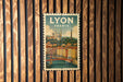 A vintage-style postage stamp from Lyon, France, featuring a cityscape with a bridge and a castle, hanging on a wooden wall.
