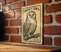 A wooden frame with a black and white illustration of a snowy owl perched on a branch, mounted on a brick wall.