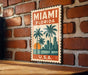 A vintage-style metal sign with the text "MIAMI FLORIDA" and a graphic of palm trees and a sunset, mounted on a brick wall.
