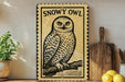 a framed illustration of a snowy owl perched on a branch, with a wooden cutting board and a potted plant in the background.
