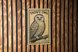 A black and white illustration of a snowy owl perched on a branch, mounted on a wooden wall.