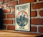 A metal sign with the text "Detroit Michigan USA" and an illustration of a city skyline is displayed on a brick wall.
