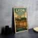 A vintage-style metal sign with the text "LYON FRANCE" and an illustration of a cityscape with a bridge, placed on a gray countertop next to some black camera lenses.