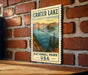 A vintage-style sign for Crater Lake National Park, featuring a scenic illustration of the lake surrounded by mountains and pine trees.